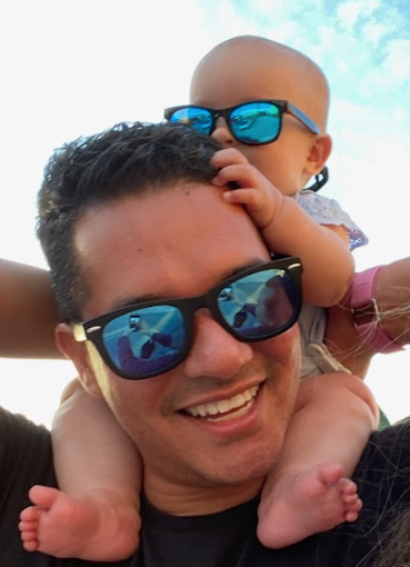 Photo of man wearing sunglasses, smiling, with his baby on his shoulders, also wearing sunglasses
