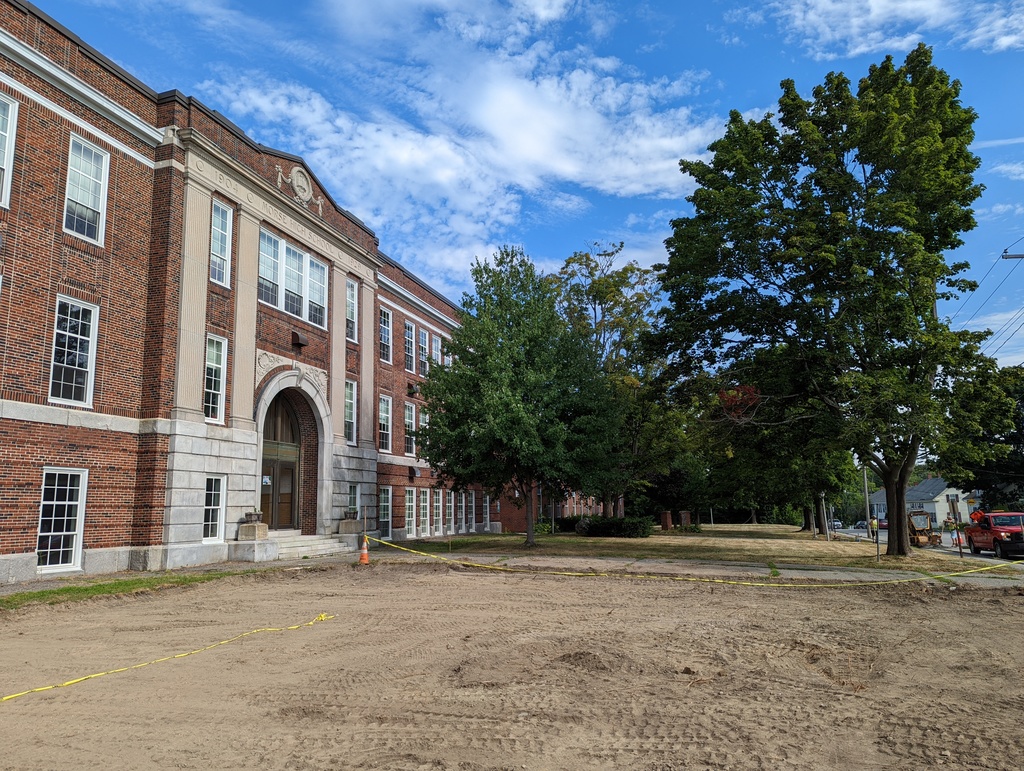 Photo shows old Morse High School on the left, with three stories, tall glass windows, and arched main entrance.  In the foreground can be seen an area of ground where topsoil has been removed and equipment will be installed.  Trees and lawn are in the background.