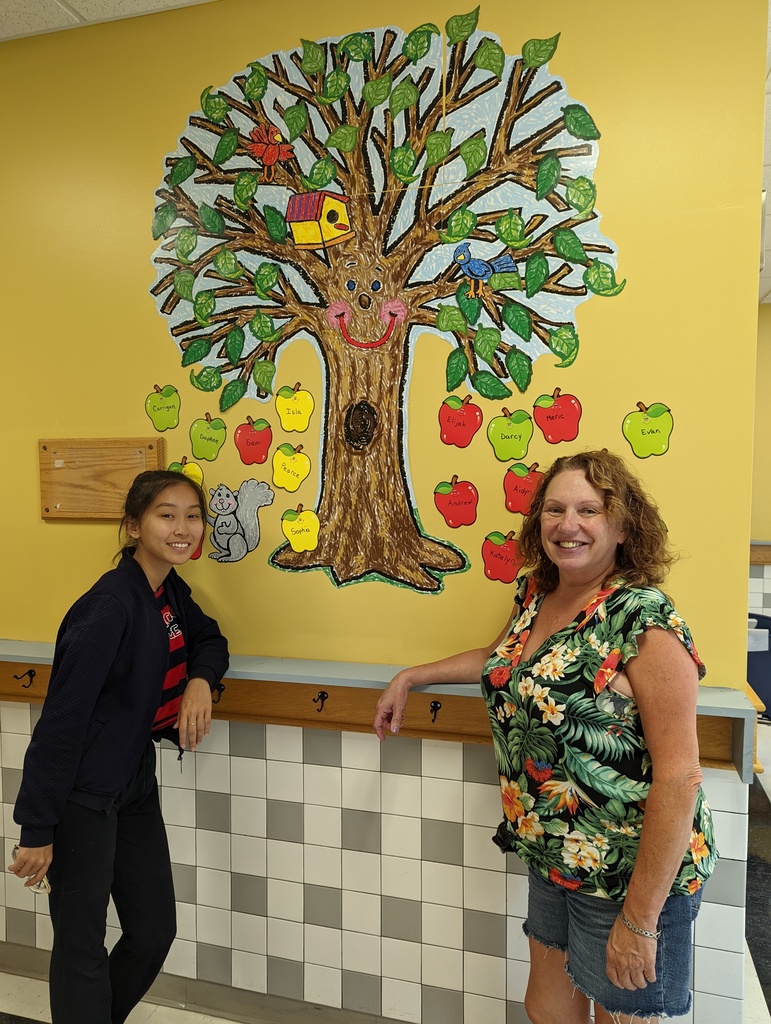 Student volunteer and teacher standing in front of a tree artwork with student names displayed in apples.