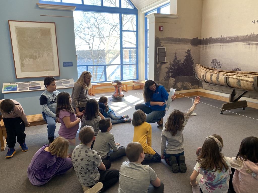 Second Grade Class of students sit and listen to a museum presenter, Native American canoe on display in the background